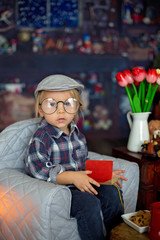 Sweet toddler boy, wearing glasses, reading a book and drinking tea with cookies
