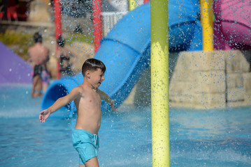 Cute European boy in blue swimming shorts having fun in aqua park he is jumping in pool making lots of splashes.