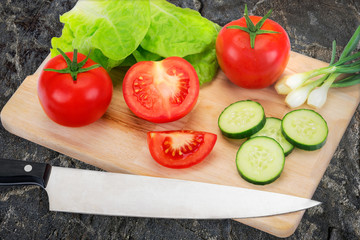 Concept of preparing a healthy salad - fresh and organic vegetables on a chopping board with a knife on a background of dark stone countertop