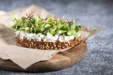 Vegetarian sandwich on a wooden Board and a piece of paper. A piece of rye bread with sesame seeds, soft cheese, sunflower sprouts, micro-greens, on a dark gray background. Health food. Close up.