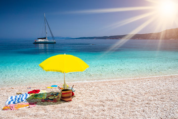 Summer concept with accessories on beach. Yellow umbrella on picturesque pebble beach straw bag, towel, Kefalonia island, Greece. Travel relax vacation concept.