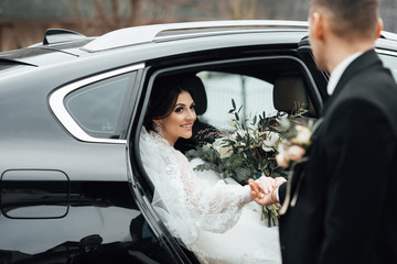 Bride with flower bouquet in car. Pretty woman wearing white wedding dress and tiara with veil. The bride gets out of the car and the groom extends a hand. Bride with wedding makeup and hairstyle