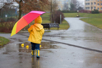 Beautiful funny blonde toddler boy with rubber ducks and colorful umbrella, jumping in puddles and...