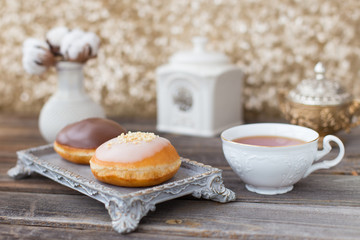Donuts made of white milk chocolate, coated with glaze and nuts. Aristocratic tea party with porcelain cup on wooden table. Decorated with vases,cotton inflorescences,white sugar bowl.Gold background