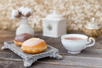 Donuts made of white milk chocolate, coated with glaze and nuts. Aristocratic tea party with porcelain cup on wooden table. Decorated with vases,cotton inflorescences,white sugar bowl.Gold background
