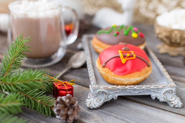 Christmas chocolate donuts with Santa Claus icing and new year tree on ceramic stand. Glass cup of cocoa with marshmallows on wooden table. Spruce branch, present, cone, spoon, sugar bowl, white deer