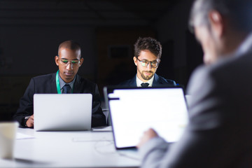 Confident people working over project in office late at night. Thoughtful business team sitting at long table with laptops. Teamwork, working late concept