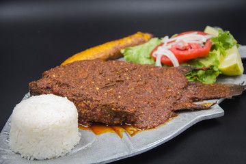 Crispy fried whole fish topped with smoky chipotle sauce, served on a unique fish-shaped metal platter with white rice and fresh salad, against a dramatic black background.