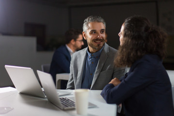 Content coworkers talking in dark office. Professional businessman and businesswoman working with laptops in dark office. Working late concept