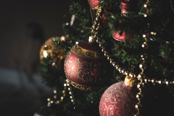 A portrait of red and gold christmas balls hanging in a tree with other christmas ornaments. The ball is red and has a gold print on it, above it there is a gold bead garland.