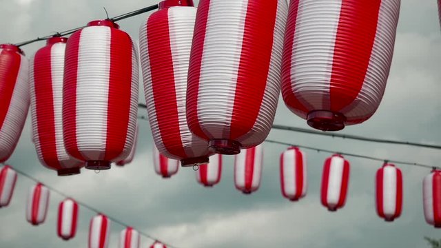 Many oriental paper red-white lanterns Chochin hanging on white cloudy blue sky background. Japanese lanterns hanging on bright sky background