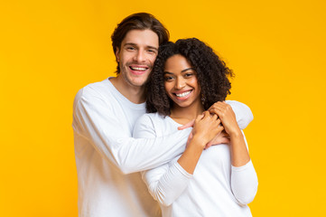Romantic multiracial couple embracing, posing together over yellow background in studio