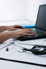 Close-up stethoscope on the doctor's desktop. In the background are hands and a laptop. The concept of a doctor in the workplace, online consultation