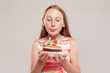 Inclusive Beauty. Girl with freckles standing isolated on grey holding birthday cake blowing candles making wish close-up