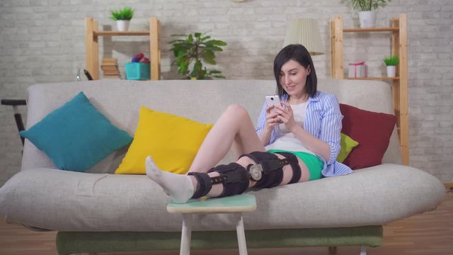 female with the phone after injury knee bend orthosis knee brace lies on the couch at home
