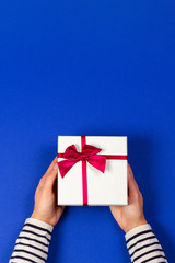 Woman hands holding present gift box tied with ribbon on navy blue background. Top view, place for text. Holiday concept