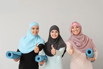 Sporty Muslim women with yoga mats showing thumb-up on light background