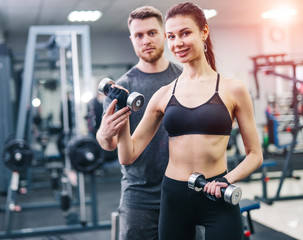 Fototapeta na wymiar Personal trainer helping woman working with heavy dumbbells indoors. Beautiful female with her fitness trainer exercising in the gym.