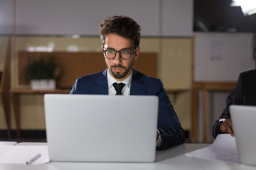 Front view of focused man in eyeglasses looking at laptop. Concentrated handsome manager sitting at table and working with laptop. Business, working late concept