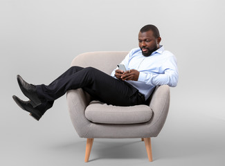 African-American man with mobile phone relaxing in armchair against grey background