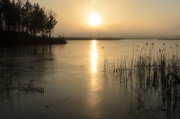 Ice of Lake at sunset with cracks and snow. Mountains beyond the lake. The sun is on the horizon. Reflection of the sun on ice.