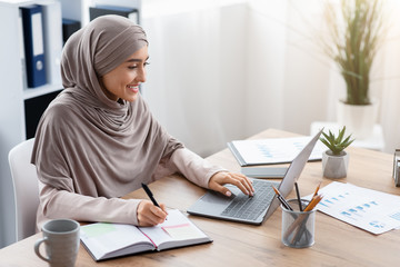 Muslim woman in hijab using laptop and taking notes in office