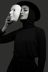 high fashion portrait of elegant woman in black dress and hat, holding theatre white mask. Studio...