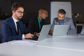 Fototapeta premium Group of confident businessmen working with laptops in office. Thoughtful young managers working together in office at night. Teamwork, working late concept