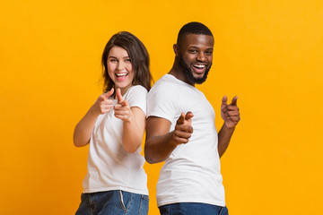Cheerful interracial couple pointing at camera with fingers over yellow background