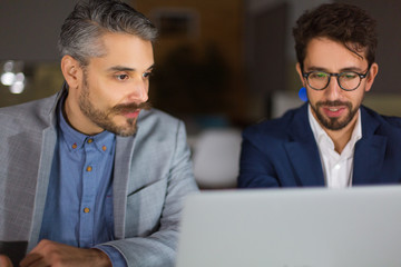 Obraz premium Two handsome businessmen looking at laptop. Front view of two business partners working with laptop. Business, working late concept