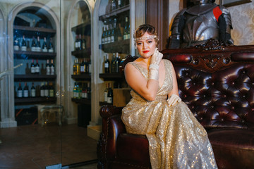 lady in a gold dress and with a diodema sits on a brown leather armchair