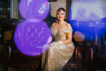 a woman from the 1920s in a gold dress sits in a leather armchair against the background are...