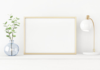 Interior poster mockup with horizontal gold metal frame on the table with plant in blue vase and lamp on empty white wall background. A4, A3 size format. 3D rendering, illustration.