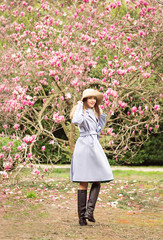 a young, beautiful woman, in a blue coat and a light hat, walks in the Park in the spring near a flowering Magnolia tree, on a Sunny day