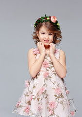 Portrait of a beautiful cut little girl in an elegant dress with a wreath of fresh flowers on her head stands, lauht and looks into the camera in the studio on a gray background.