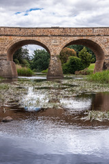 Fototapeta na wymiar Richmond, Tasmania, Australia - December 13, 2009: Aong water line closeup portrait shot of part of brown stone historic bridge over coal river reflected in water with reed. Some blue cloudscape