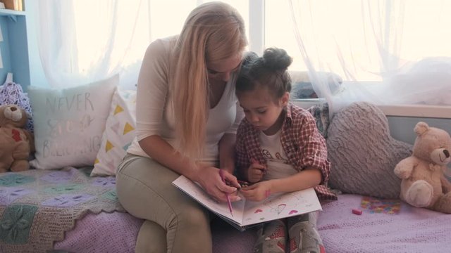 Young woman and her little daughter spent time together, they paint a coloring book with colored pencils
