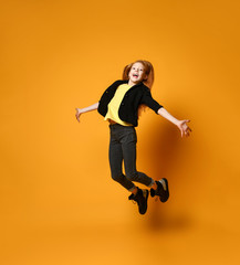 Fototapeta na wymiar Teen female in black jacket, pants and boots, yellow t-shirt. She jumping up with spreaded hands, posing on orange background.