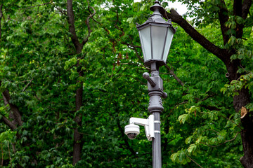 Iron street lamp with video surveillance camera in the park on a background of green trees.