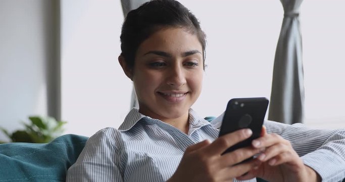 Smiling indian young woman using smartphone apps sit on sofa
