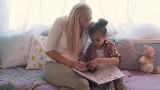 Mother and her little daughter spent time together, talking and they paint a coloring book with colored pencils