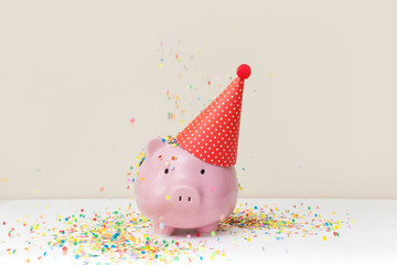 Pink piggy bank with birthday cap and color confetti on beige background