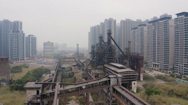 Slow dolly in on the abandoned steel mill factory with blast furnaces structures in between newly built apartment blocks on a cloudy day in Guangzhou, China, Aerial shot.