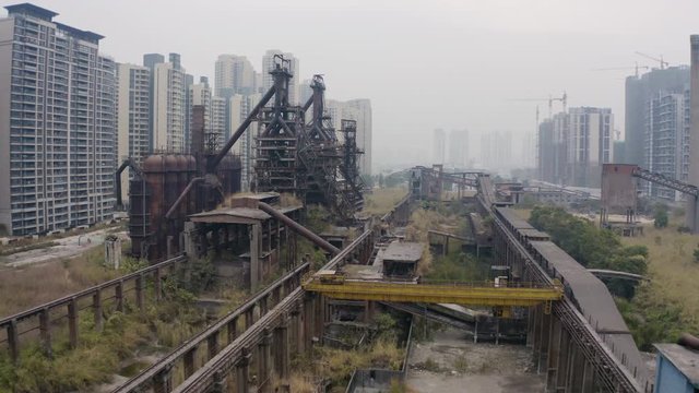 Fly over the abandoned steel mill factory with blast furnaces structures in between newly built apartment blocks on a cloudy day in Guangzhou, China, Aerial shot.