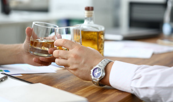 Close-up of businessmen hands clinking glasses with whiskey. Colleagues celebrating signing contract. Partners relaxing and drinking scotch after busy day. Business negotiations concept
