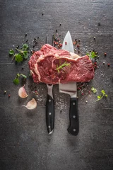 Tuinposter Raw beef steak with fork and knife salt pepper and herbs on dark concrete background © weyo