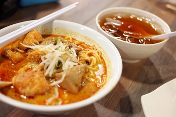 Delicious bowl of curry laksa with noodles, tofu puffs, and chicken, served with a side drink.