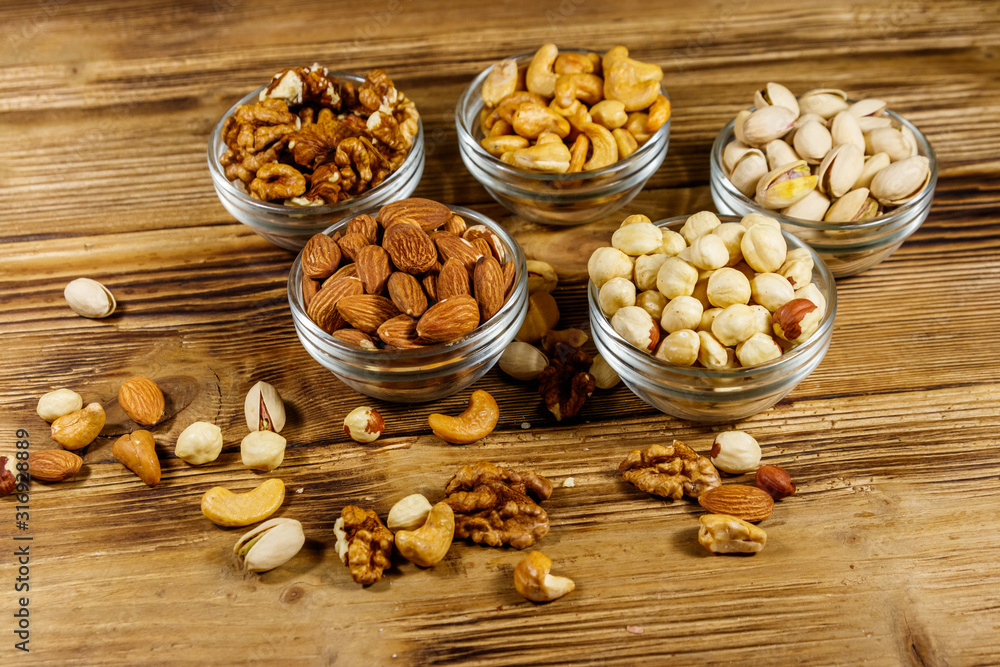 Poster Assortment of nuts on wooden table. Almond, hazelnut, pistachio, walnut and cashew in glass bowls. Healthy eating concept