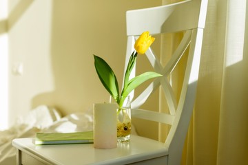 Yellow tulip flower in glass with water on white chair in bedroom interior