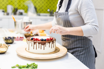 Confectioner decorates with berries a biscuit cake with white cream and chocolate. Cake stands on a wooden stand on a white table. The concept of homemade pastry, cooking cakes.